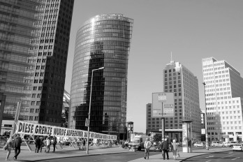 Das Transparent zum 3. Oktober 2015 auf dem Potsdamer Platz auf der Grenze zwischen Ost- und Westberlin – zwischen „Deutsche-Bahn-Tower” (mittig), dem nach dem Altnazi Otto Beis­heim benannten „Beisheim Center” (die zwei Gebäude rechts), Polizeifahrzeugen und „Mauer”-Gedenksteinen recht eingequetscht